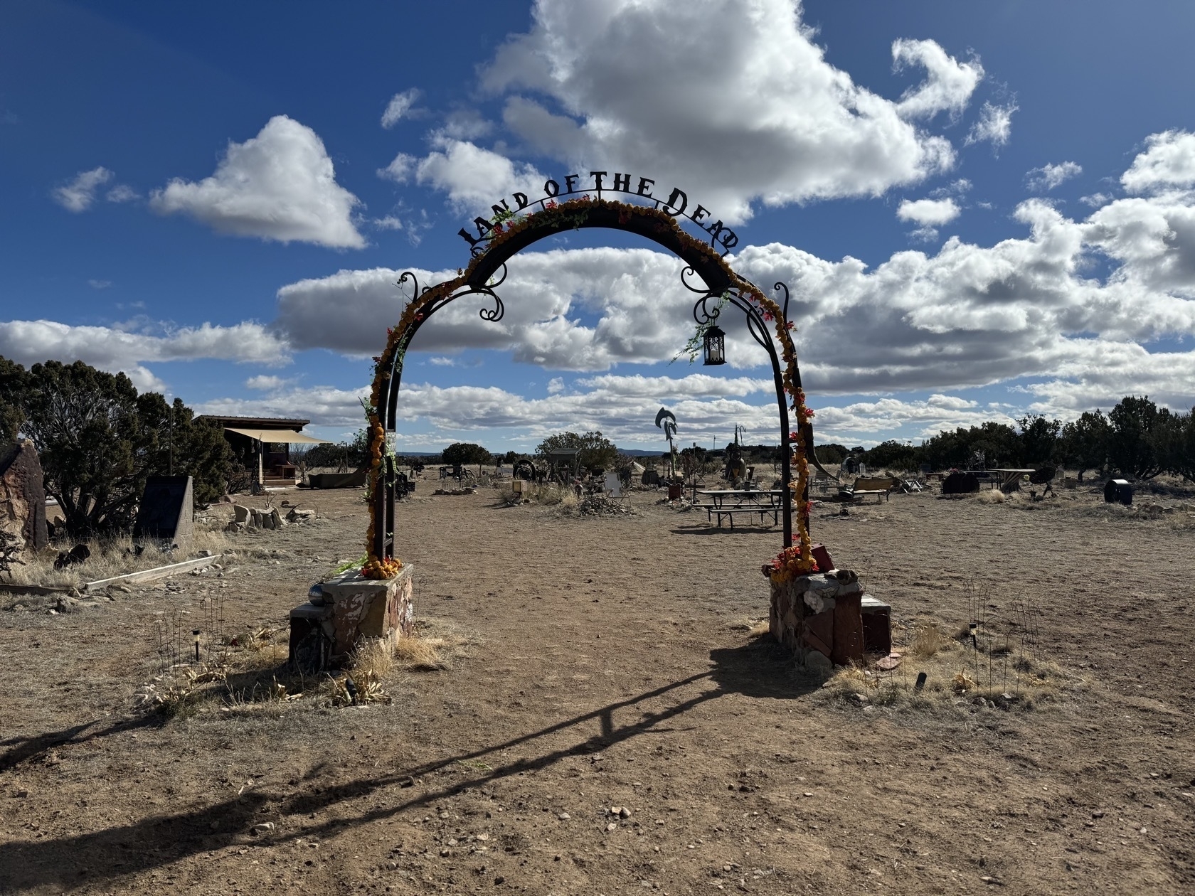 Arched metal gate centered in frame with scrubby desert landscape in foreground and bright blue sky above. The gate has flowers hanging down the sides and reads Land of the Dead across the top. In the background are graves, memorials, and assorted seating areas and decorations.