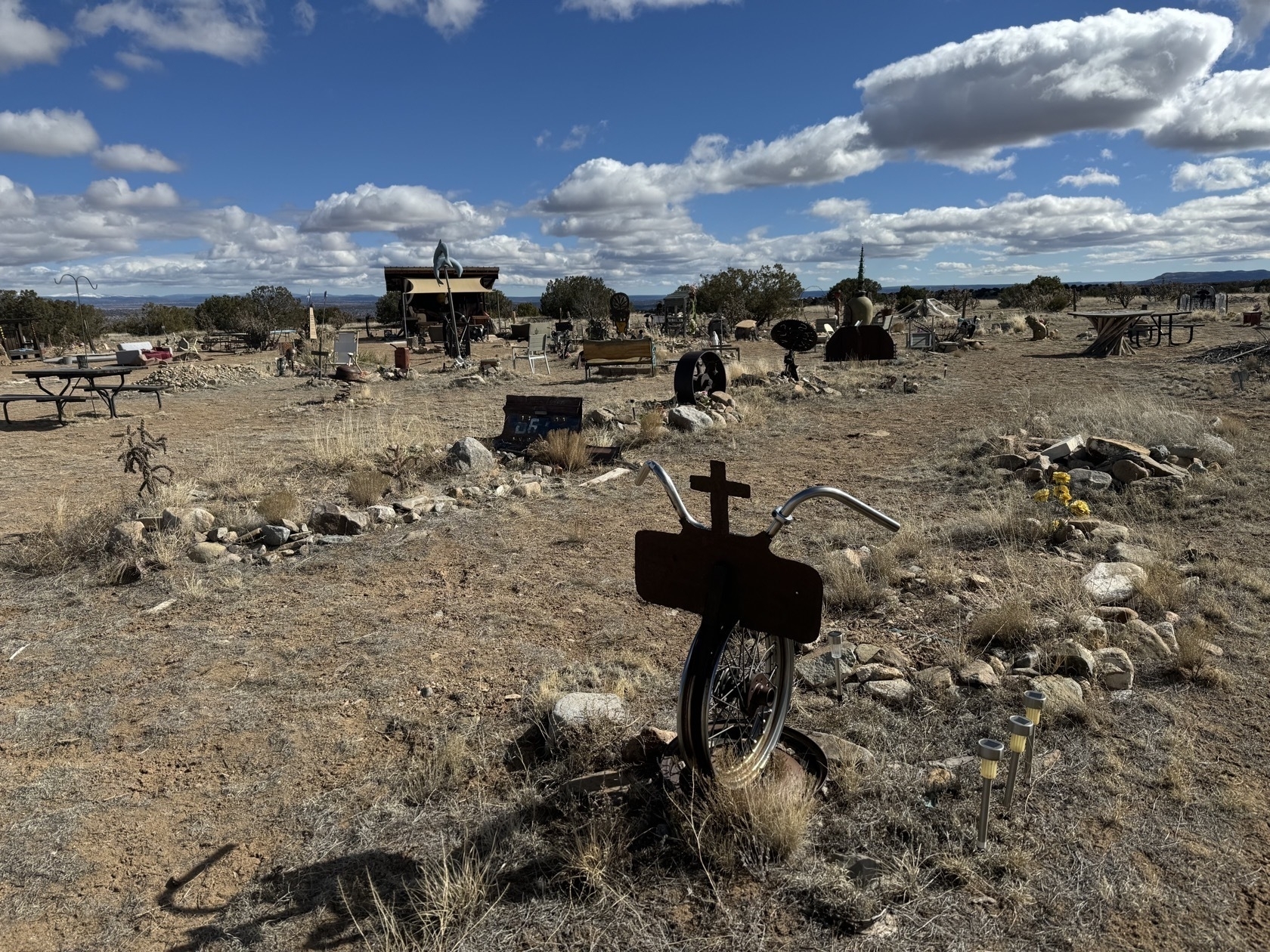 In the foreground a memorial is made from a bicycle wheel and handlebars with a cross in the middle. The background shows a diversity of non-traditional memorials and headstones.